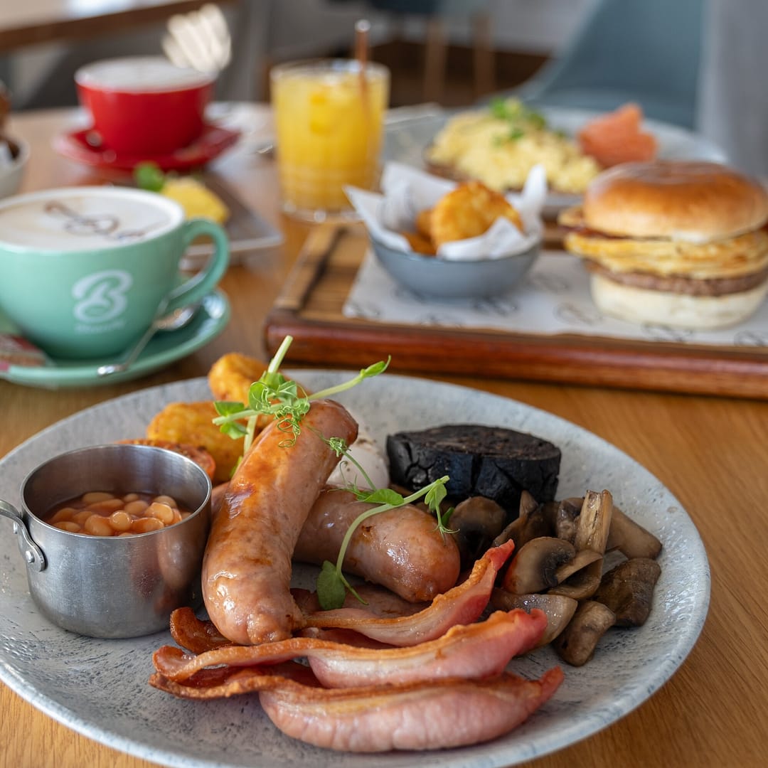 Full breakfast with sausages, bacon, beans and mushrooms, with coffee and brunch dishes behind